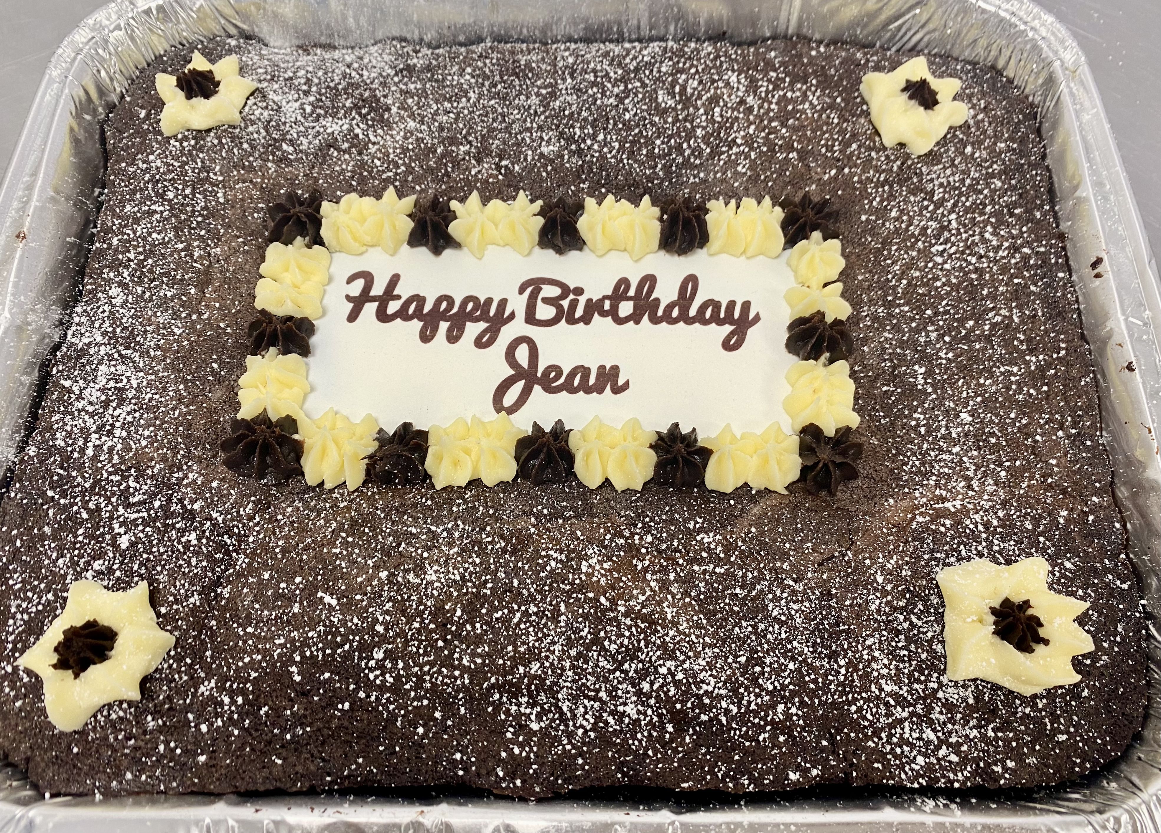 Chocolate brownies in a foil tray, boasting a rich, chocolatey flavor and decorated with powdered sugar. It has a white plaque in the center with the message 'Happy Birthday Jean' written in brown icing, bordered by yellow and brown frosting flowers. The corners feature additional yellow frosting flowers.